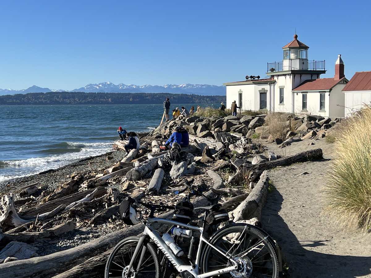 bikerumor pic of the day a bicycle leans against some logs washed up on shore, there is a white light house building in the background and a city on the horizon.