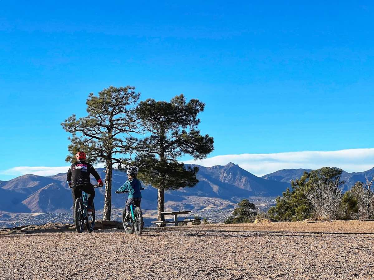 bikerumor pic of the day two people, one adult and one child sit atop mountain bikes near two pine trees overlooking a mountain range, it is bright, the sun is low in the deep blue sky and there are whispy clouds along the the tops of the mountain ridge in the distance.