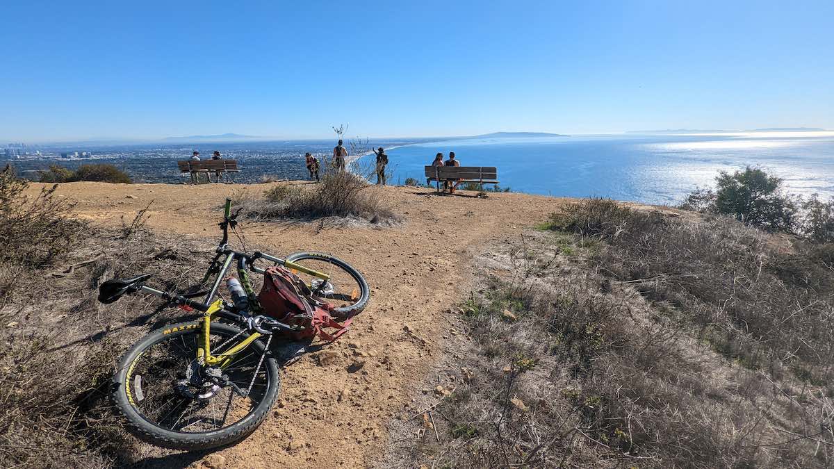 bikerumor pic of the day a bike with packs leans on its side at a mountain overlook above the sea on a sunny day the area is dirt with small scrub brush surrounding, the sky is clear and blue