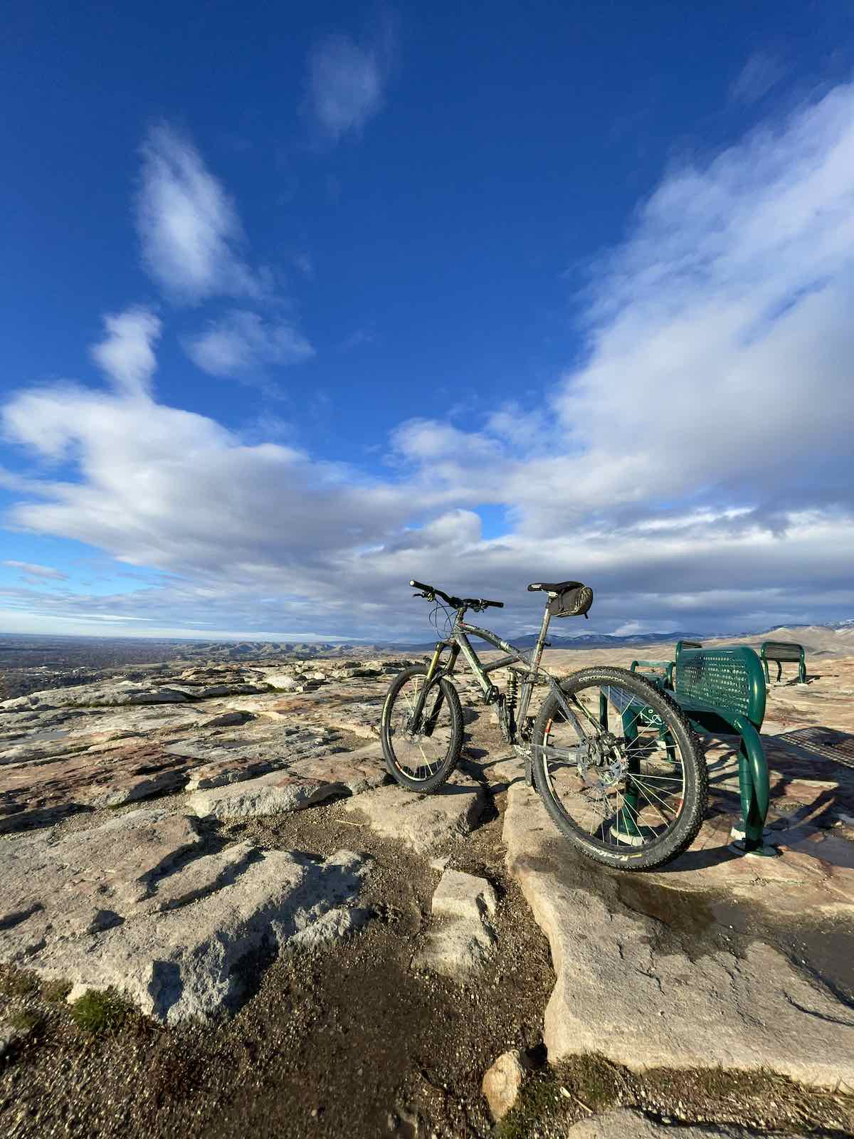 bikerumor pic of the day a mountain bike leans against a green metal bench at the top of a rocky mesa, the perspective is steep as the clouds in the sky seem to be very low and angular.