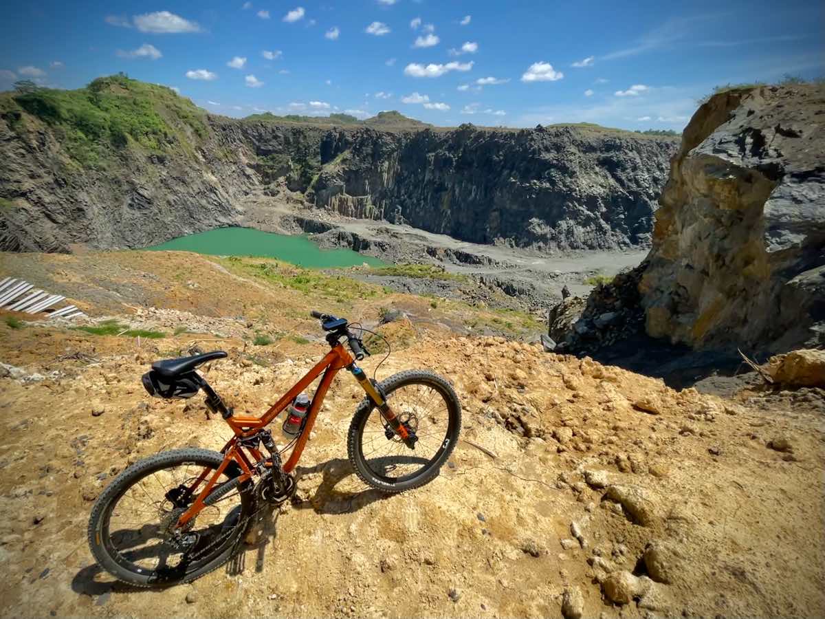 bikerumor pic of the day a bicycle is on a dirt clearing overlooking a small green pond that is surrounded by some rocky mountains, the sky is bright and blue and is dotted with small white puffs of clouds.
