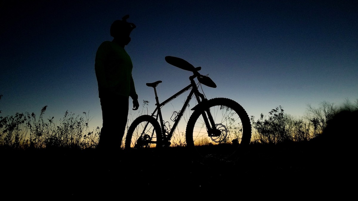 bikerumor pic of the day a cyclist stands next to their bike in profile as the sky behind is lit from the sun setting in yellow and dark blue.