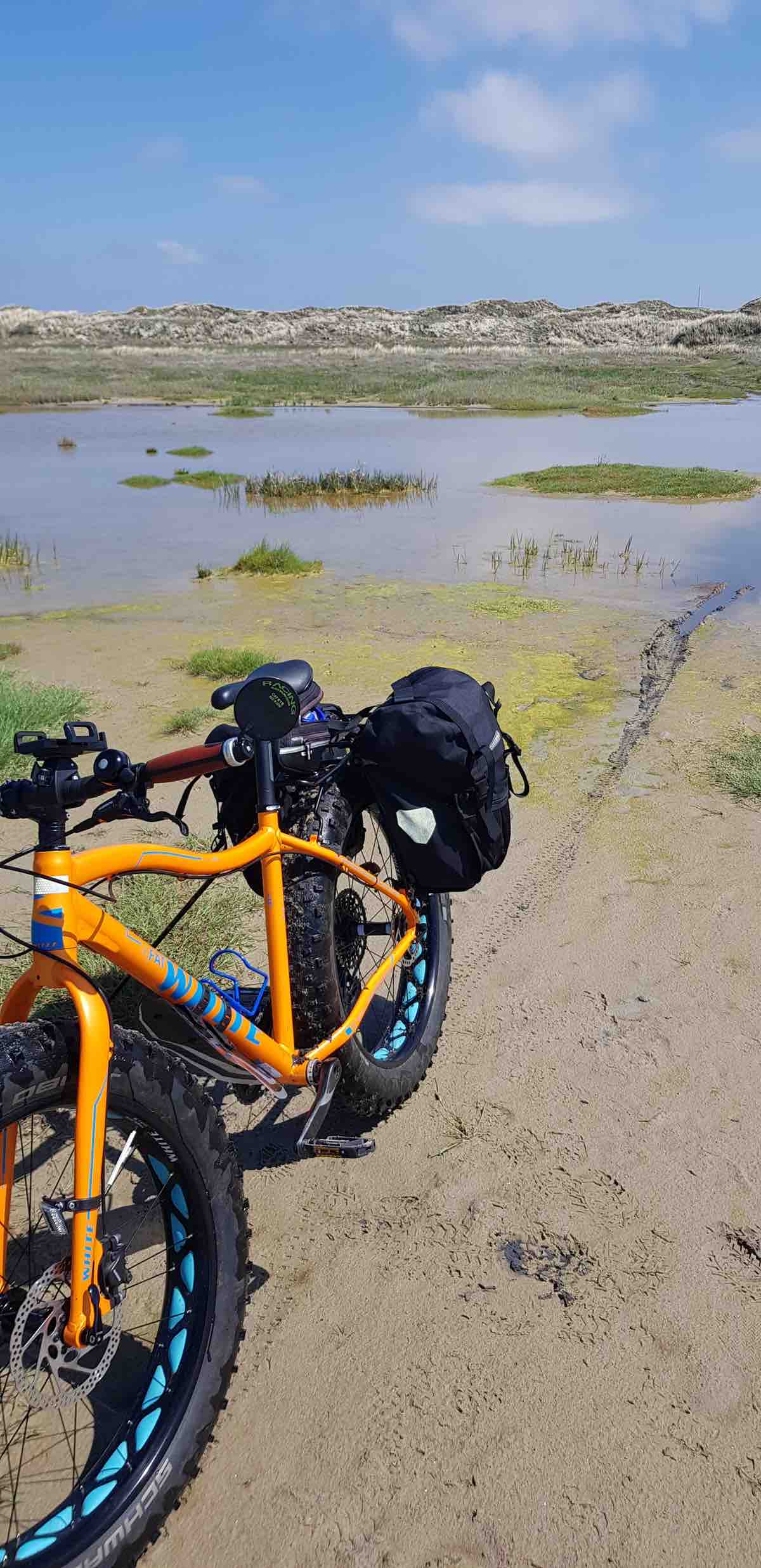 bikerumor pic of the day a fat bike with panniers is on the edge of a body of water with small hills in the distance and a light blue sky with whips of clouds.