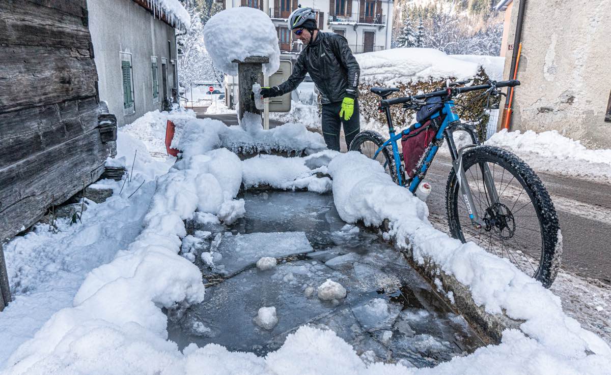 bikerumor pic of the day a cyclist stands at a water fountain that is mostly frozen and has clumps of snow around it with their water bottle and bicycle leaning against the fountain along a snowy street in chamonix france.