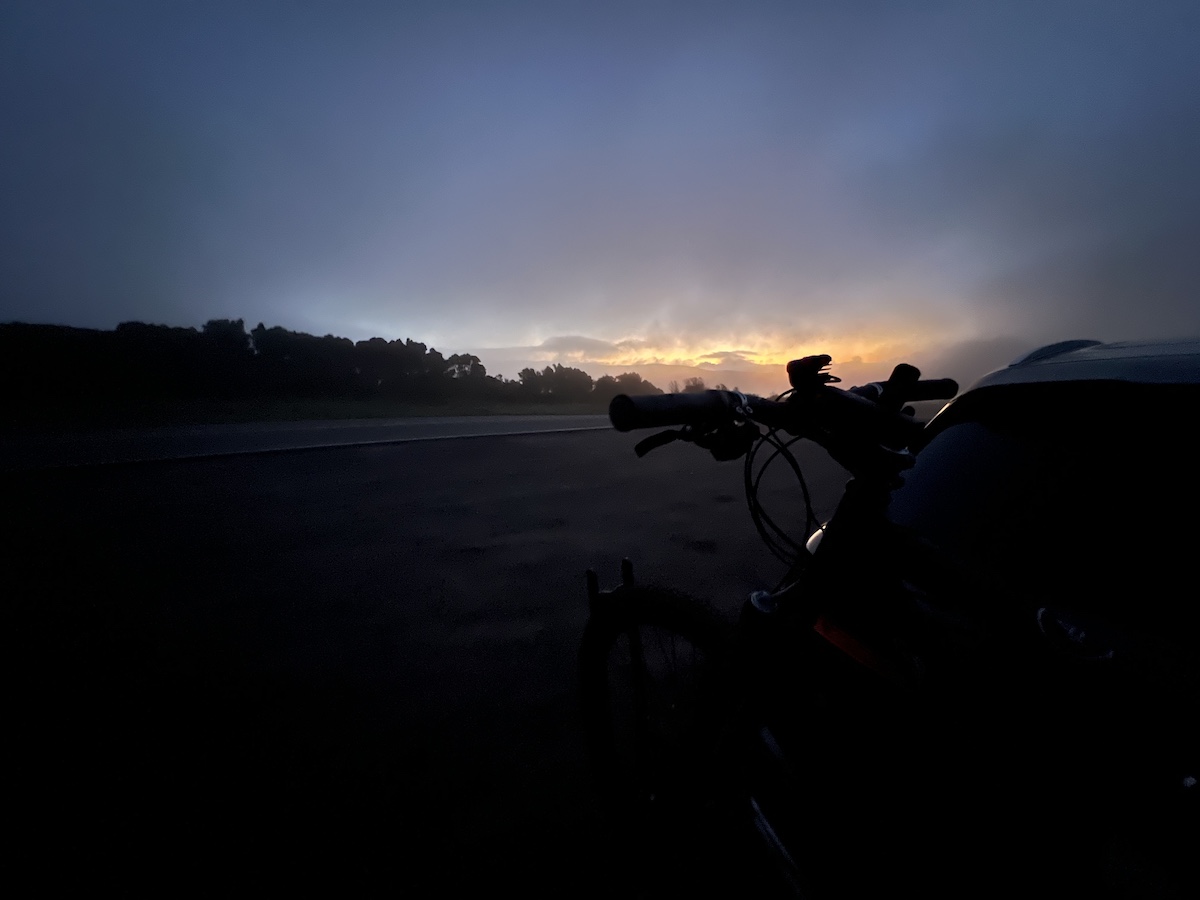 bikerumor pic of the day a close up photo of a bike looking at it's handlebars with the horizon and the sun barely peeking out in the distance, the photo is overall very dark except for the outline of the handlebar.