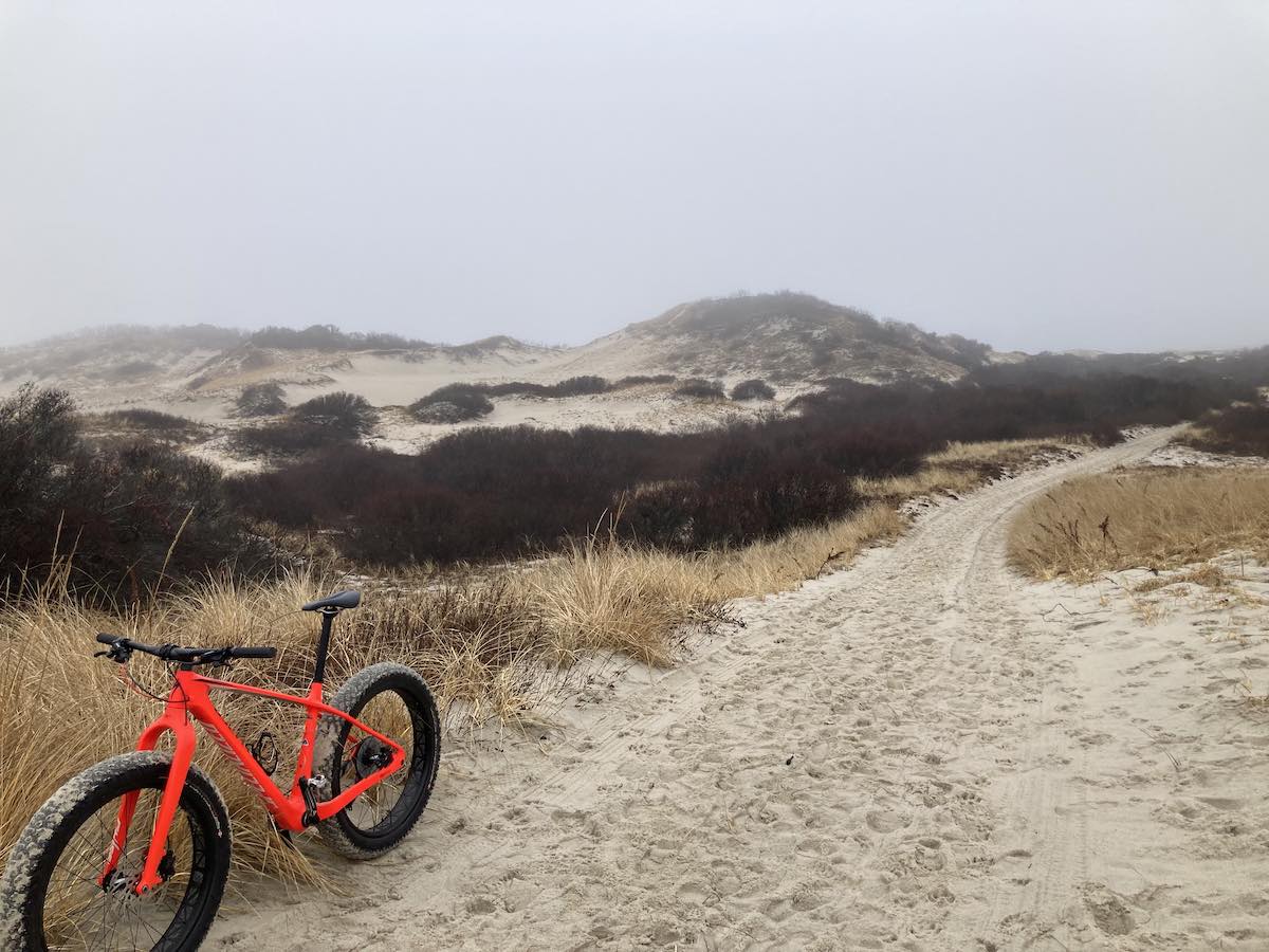 bikerumor pic of the day a red bicycle is on the side of sandy trail among grasses leading toward a sandy hill, the sky is foggy and grey and covers the top of the hill in the background