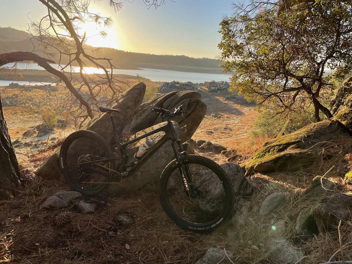 bikerumor pic of the day a mountain bike leans against a rock outcropping on the side of a trail near a body of water, the sun is low and golden over the small hill on the other side of the water.