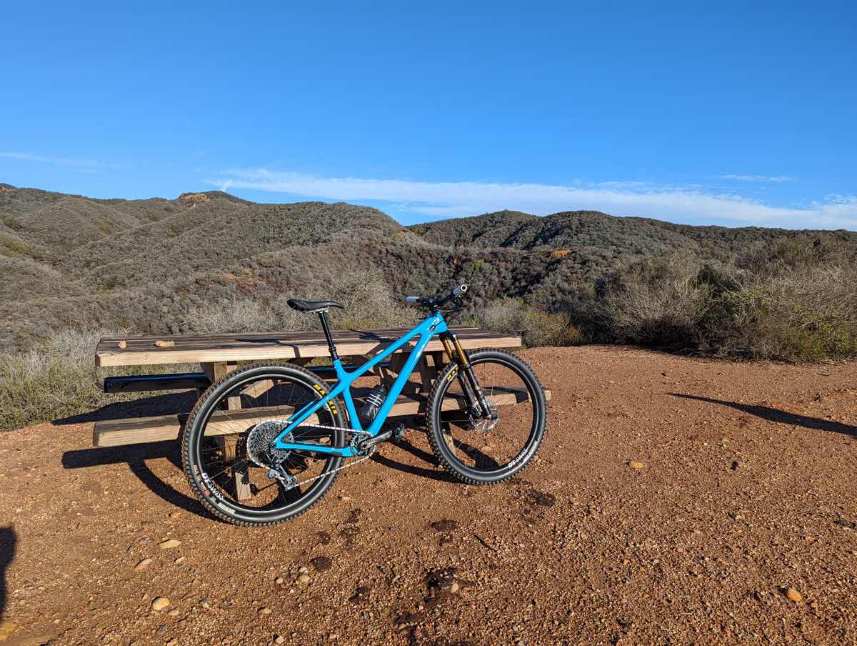 bikerumor pic of the day a mountain bike leans against a picnic table on a dirt mesa overlooking rolling hills covered in brush, the sun is bright and the sky is clear and blue.