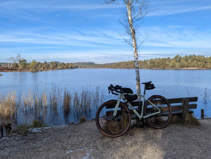 bikerumor pic of the day a canyon gravel bike leans against a wood bench next to a small lake bordered by trees, the sky is blue with whispy clouds.
