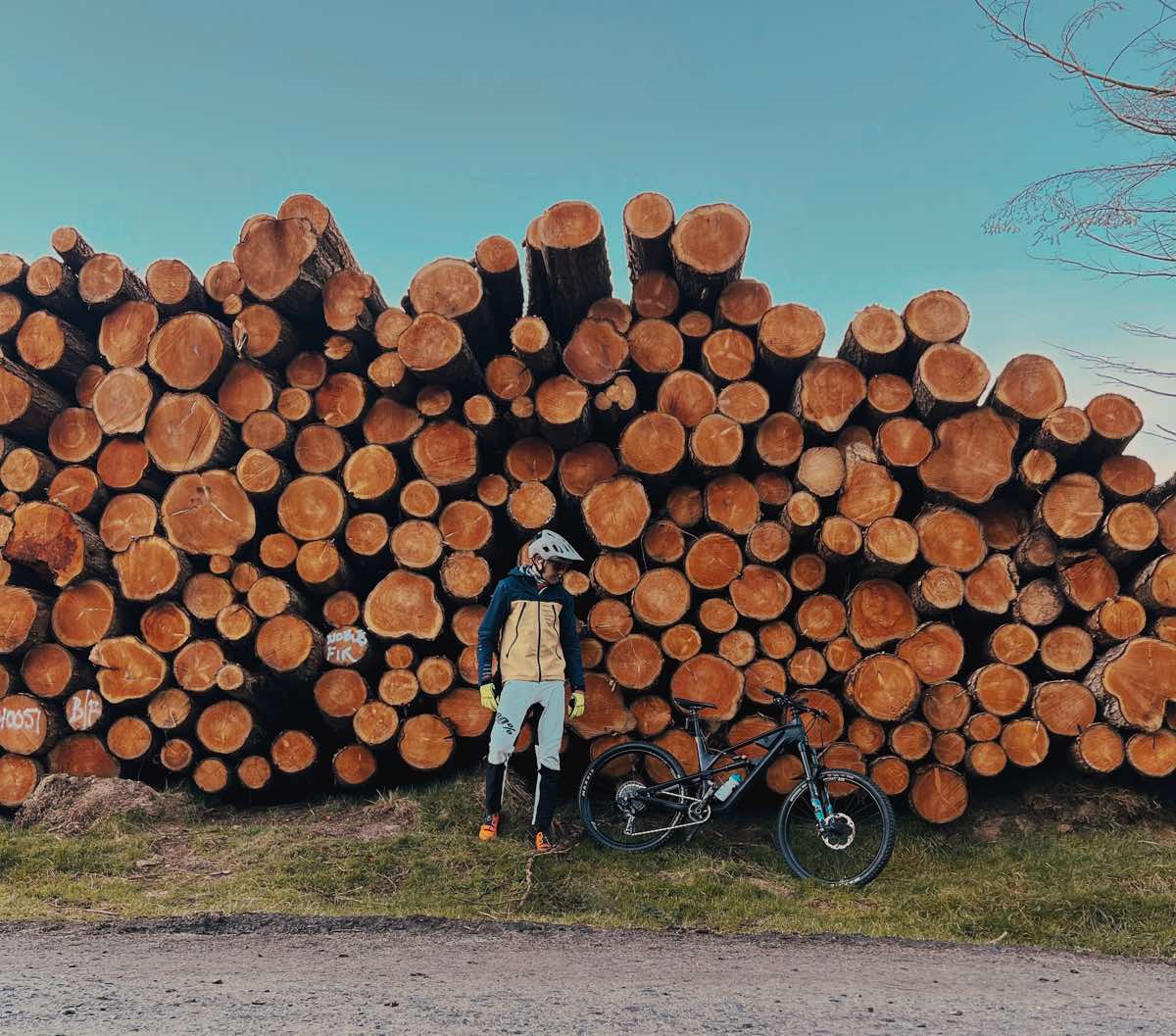 bikerumor pic of the day a cyclist stands in front of a pile of logs with their mountain bike, they are along a dirt road the the sky is clear and blue above the pile of logs.