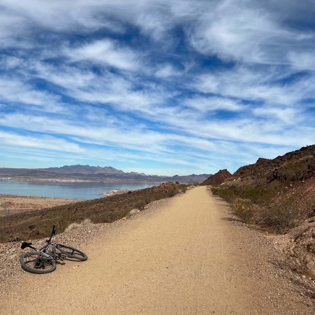 bikerumor pic of the day a mountain bike lies on its side along a wide dirt trail on the side of a long low mountain, the sky is large and covered in cotton candy like clouds with the blue sky behind peeking out