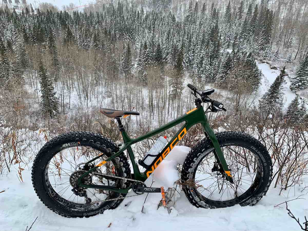bikerumor pic of the day a norco fat tire bicycle is in the snow along the edge of a path overlooking pine trees and other leafless trees down below.