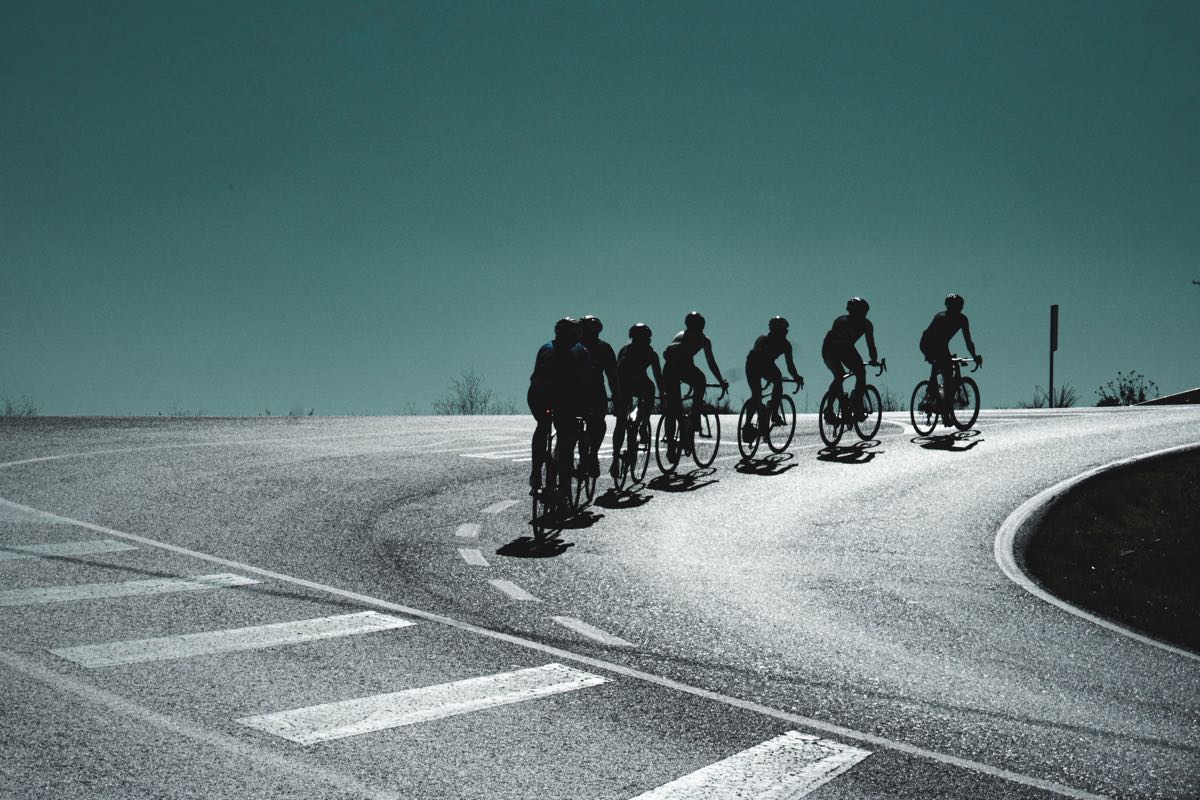 bikerumor pic of the day a group of cyclists in single file line in profile riding up a curved in the road, the sun shines on the pavement and the angle of the photo has only metallic green sky beyond the road.