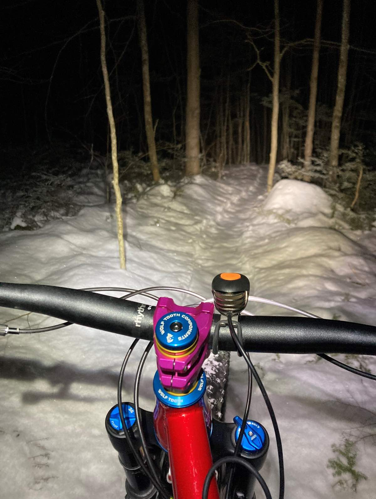 bikerumor pic of the day a view of a snow covered trail at night with the woods surrounding in darkness as a light shines on the trail over the handlebar and colorful cockpit of a mountain bike.