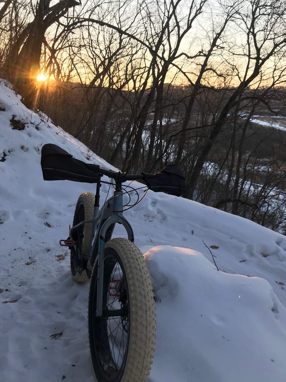 bikerumor pic of the day a fat bike with white tires and bar mitts is on the side of a sloped trail covered in snow, the sun is setting behind some bare trees over the hills and the sky is light before the sun sets.