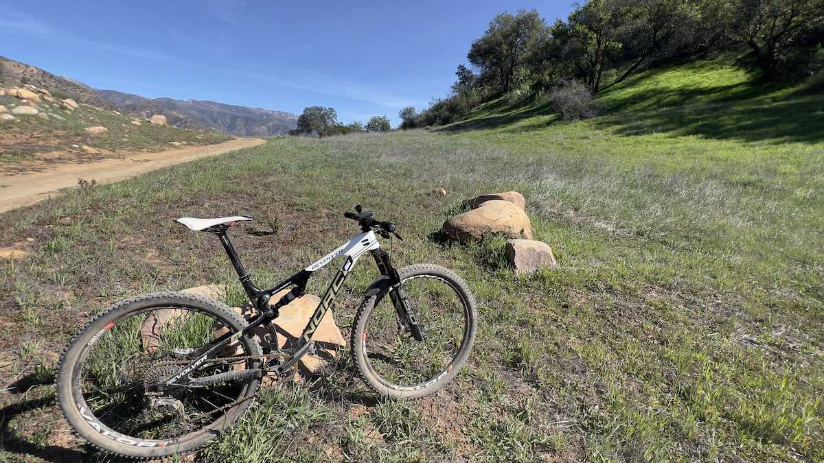 bikerumor pic of the day a mountain bike is leaned against a small rock on the grassy edge of a dirt trail, there are fluffy green trees in the distance, the sun is bright and the sky is clear blue.