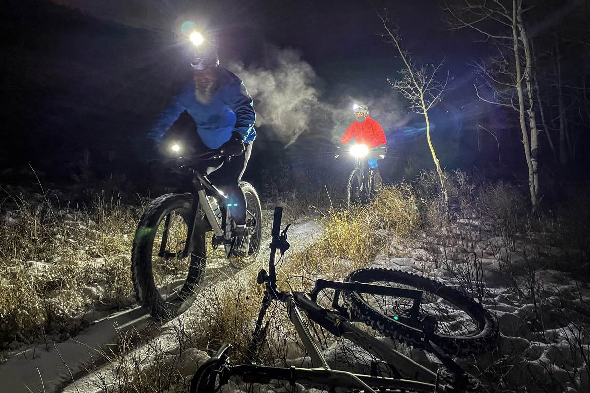bikerumor pic of the day two fat bike cyclists riding towards the left of the camera at night with their lights on a dirt trail with tall grasses on either side and steam coming off their bodies because it is so cold outside.