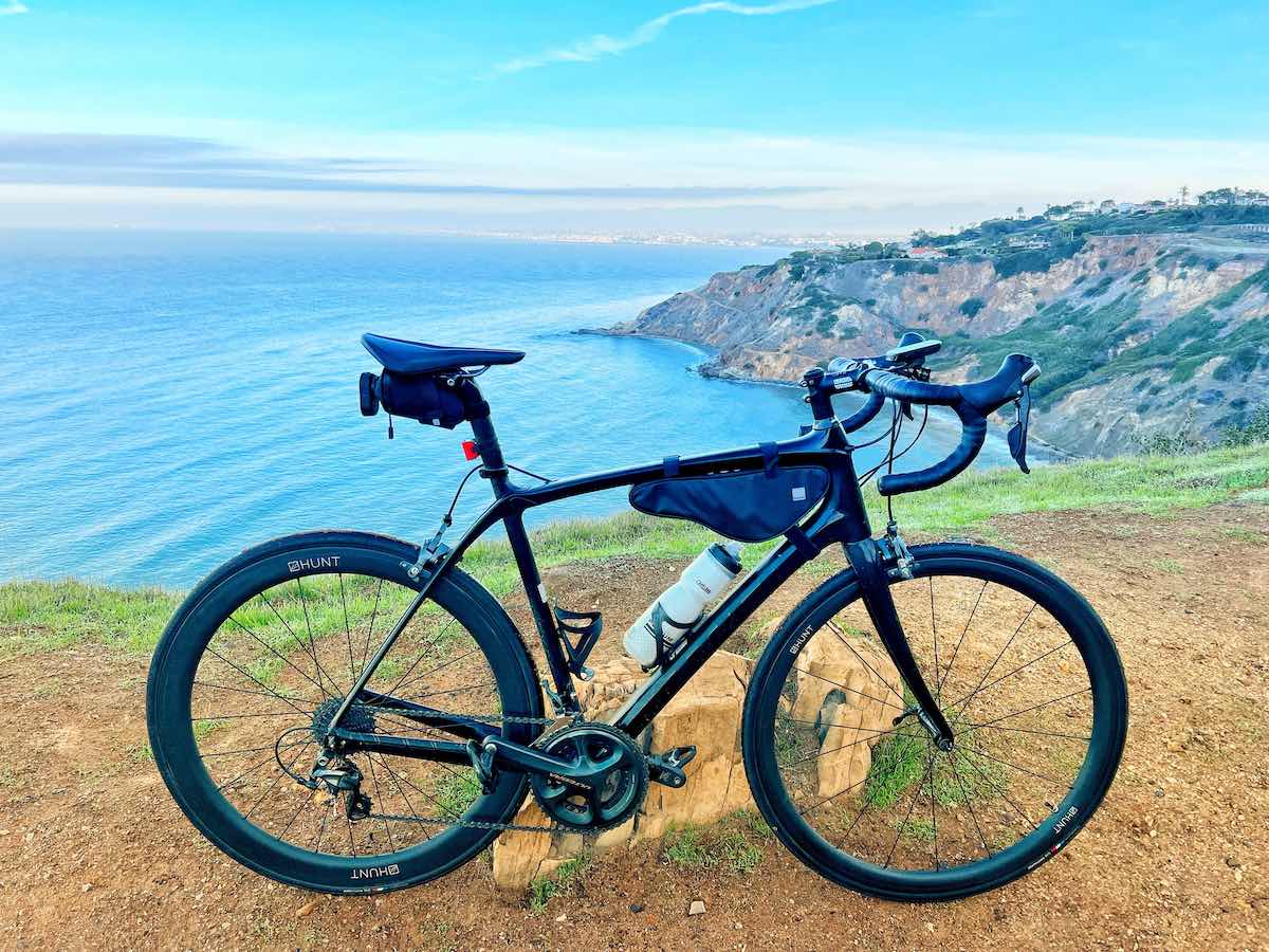 bikerumor pic of the day a road bicycle is positioned on top of a hill overlooking the ocean, the bicycle is on dirt with some grass nearby, the land below contains a small town, the sky is clear with some clouds on the horizon and the water is reflecting the bright sky