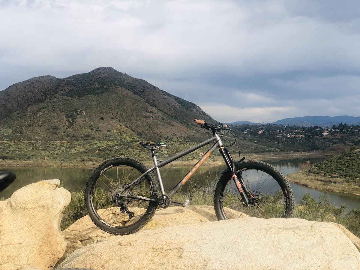 bikerumor pic of the day hardtail mountain bike is positioned on a large rock outcropping looking out over a lake with a small mountain on the other side, the sky is cloudy but the day is bright.