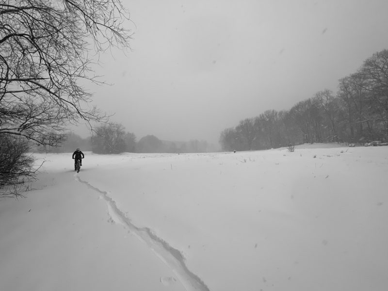 bikerumor pic of the day an open field covered in snow a lone bike rider has made a trail along the edge of the field near a bank of trees, the sky is heavy with clouds and everything is grey and white from the weather.