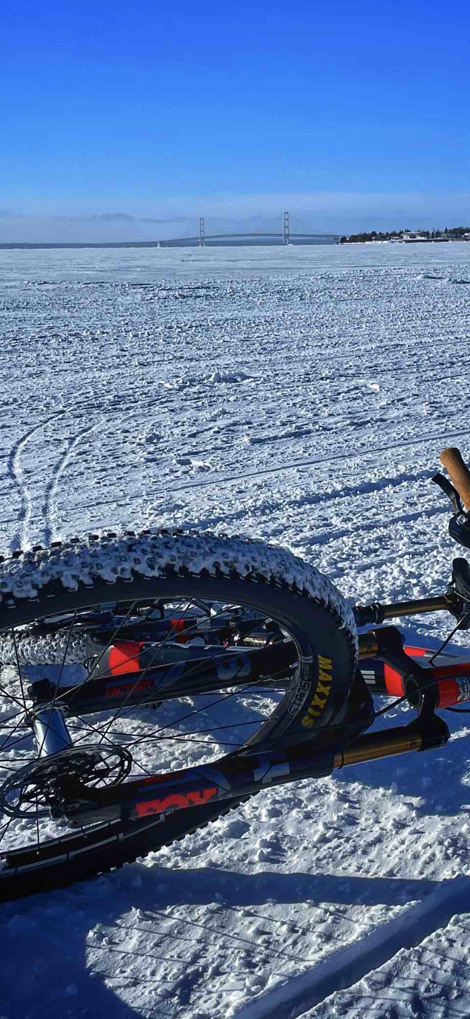 bikerumor pic of the day a mountain bike is lying on its side on an ice and snow covered expanse of water there is a bridge in the distance and the sky is clear and bright