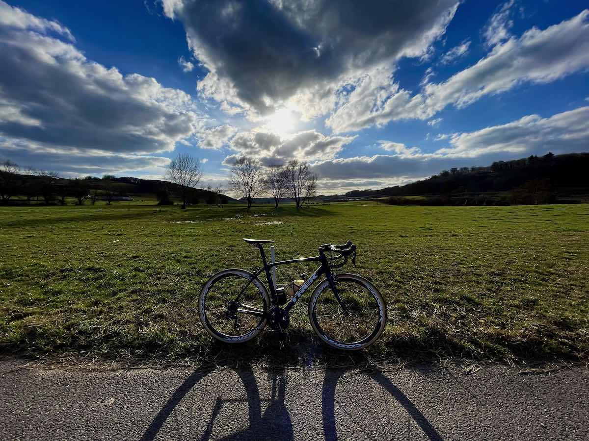 bikerumor pic of the day a bicycle is posed on the side of a road, there is a green grass field beyond it, low mountains covered in trees, the sky is bright blue with large fluffy clouds and the sun is peeking out in the middle of the photo.