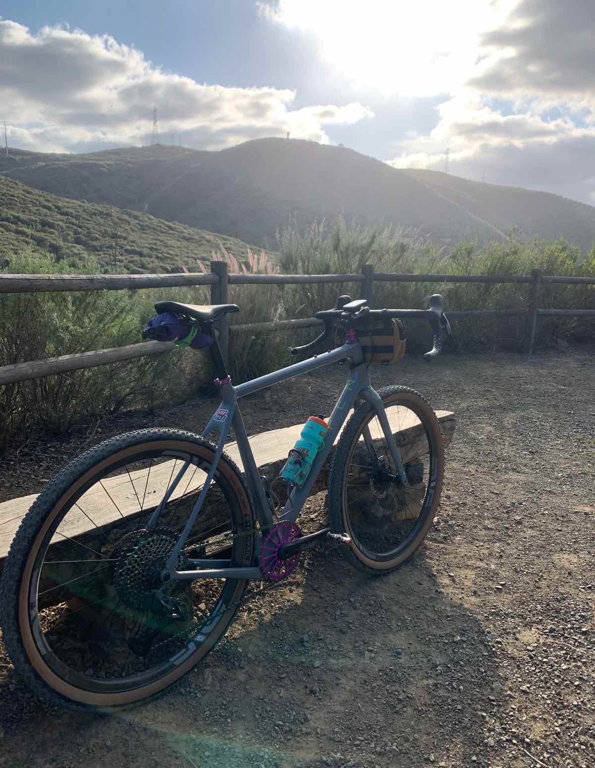 bikerumor pic of the day a mountain bike leans against a bench along a gravel trail that is enclosed with a wood railing, beyond is a green hillside with the sun peeking out through the clouds towards the viewer, it is the bright hour of the day before the sun sets.