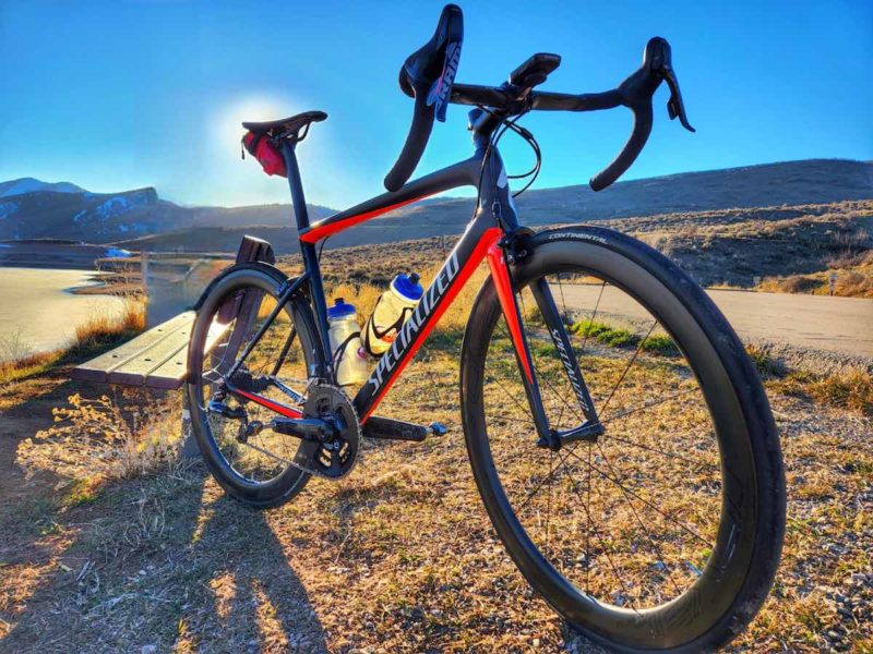 bikerumor pic of the day a specialized gravel bicycle in the grass alongside a paved trail, the sun is low in the sky over the mountains and the sky is clear and bright blue.