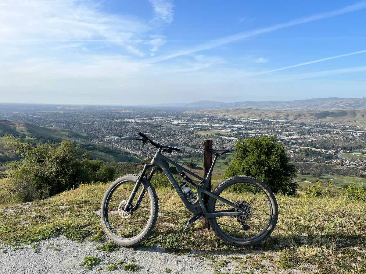 bikerumor pic of the day a mountain bike leans against a wood marker at the top of a mountain overlooking a city in a vast flat valley, the sky is hazy with clouds at the horizon and clear and blue above.
