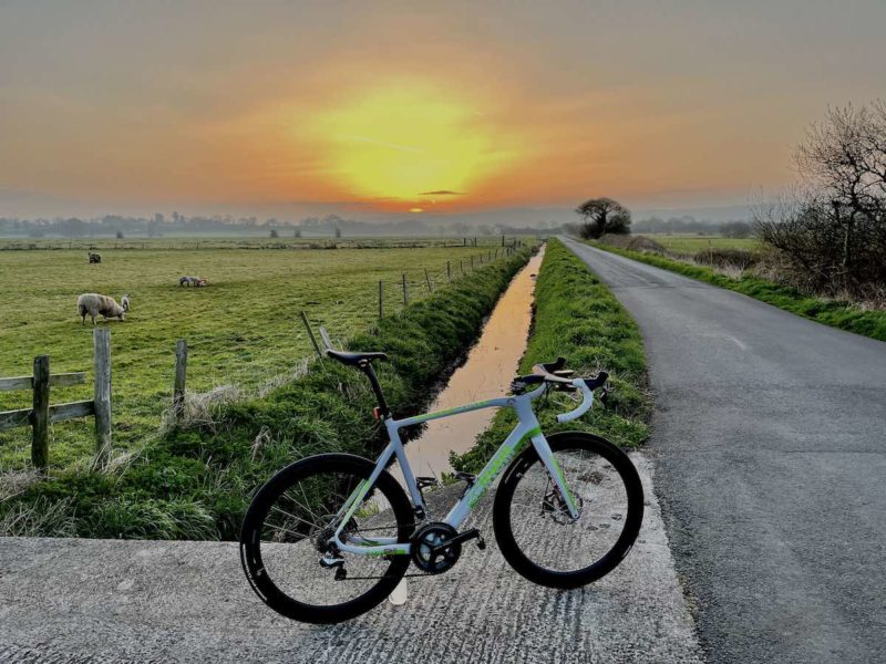 bikerumor pic of the day a bicycle is on a bit of road that goes over a culvert next to a road, there are fields on the other side of the culvert as far as the eye can see the sun is rising low on the horizon creating a hazy orange glow