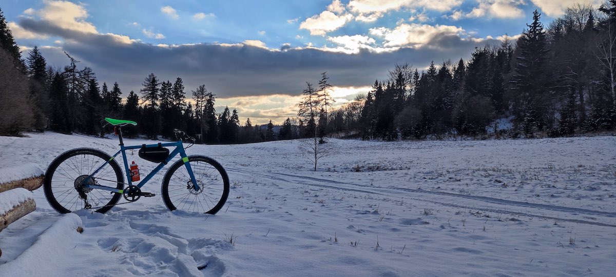 bikerumor pic of the day a photo of a bike in a field covered in snow and surrounded by a pine forest, the sky is glowing orange and blue among the clouds