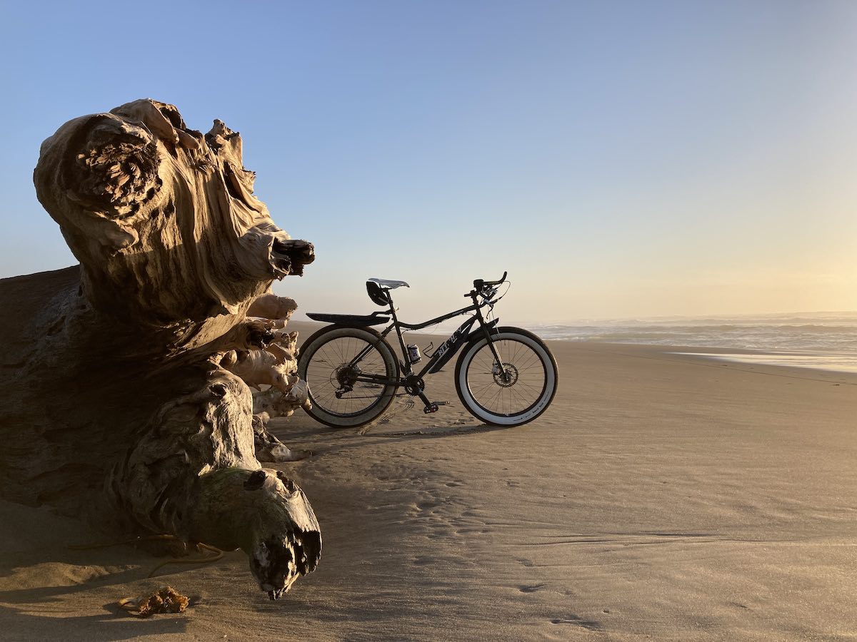 bikerumor pic of the day a ritchey fat bike leans against a giant weathered tree stump on the beach as the sun sets, there are no clouds in the sky and the horizon is bright