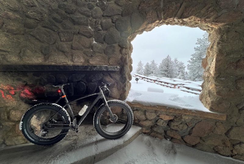 bikerumor pic of the day a fat bike is inside a stone building by a hearth with an open window, outside the window snow is covering the ground and trees nearby, the sky is cloudy and it looks like it could be snowing.