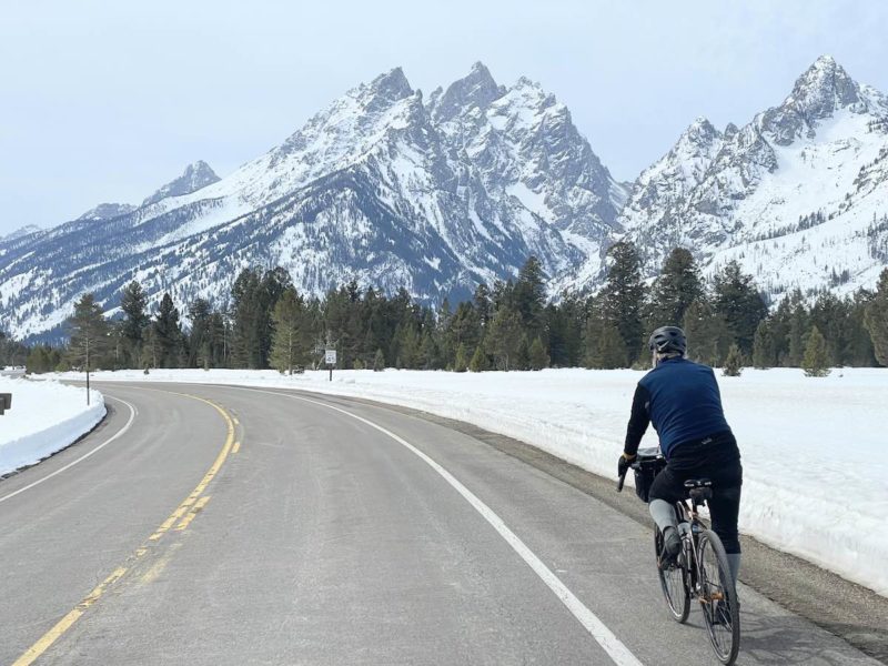 bikerumor pic of the day grand teton national park a cyclist rides on the road that is clear of cars there is snow on the side of the road and tall pointy mountains in the distance with a base of pine trees surrounding, the sky is grey.