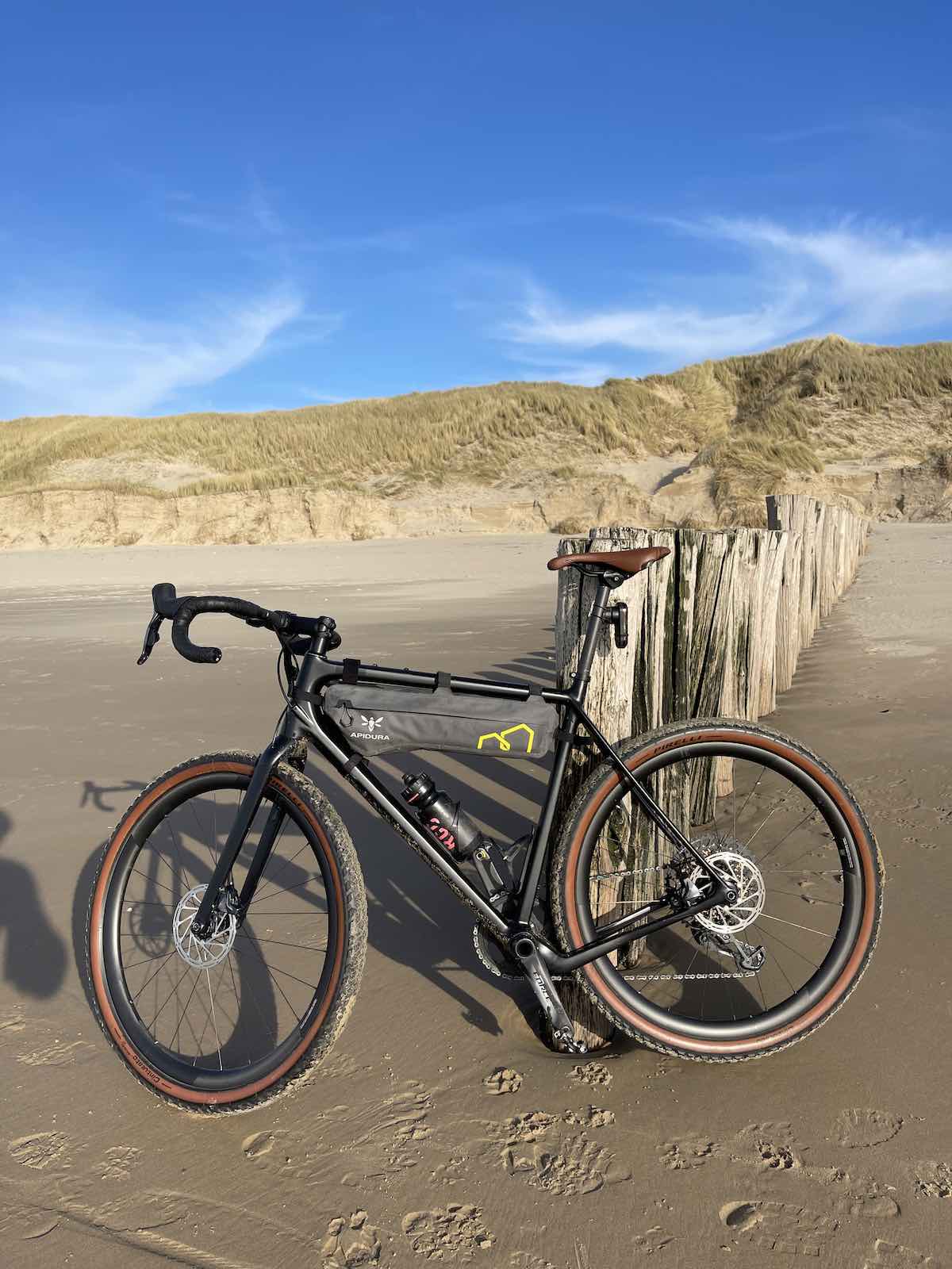 bikerumor pic of the day a bicycle leans against short wood pilings on a beach, grass covered dunes are in the background and there a a few whispy clouds in the bright blue sky.