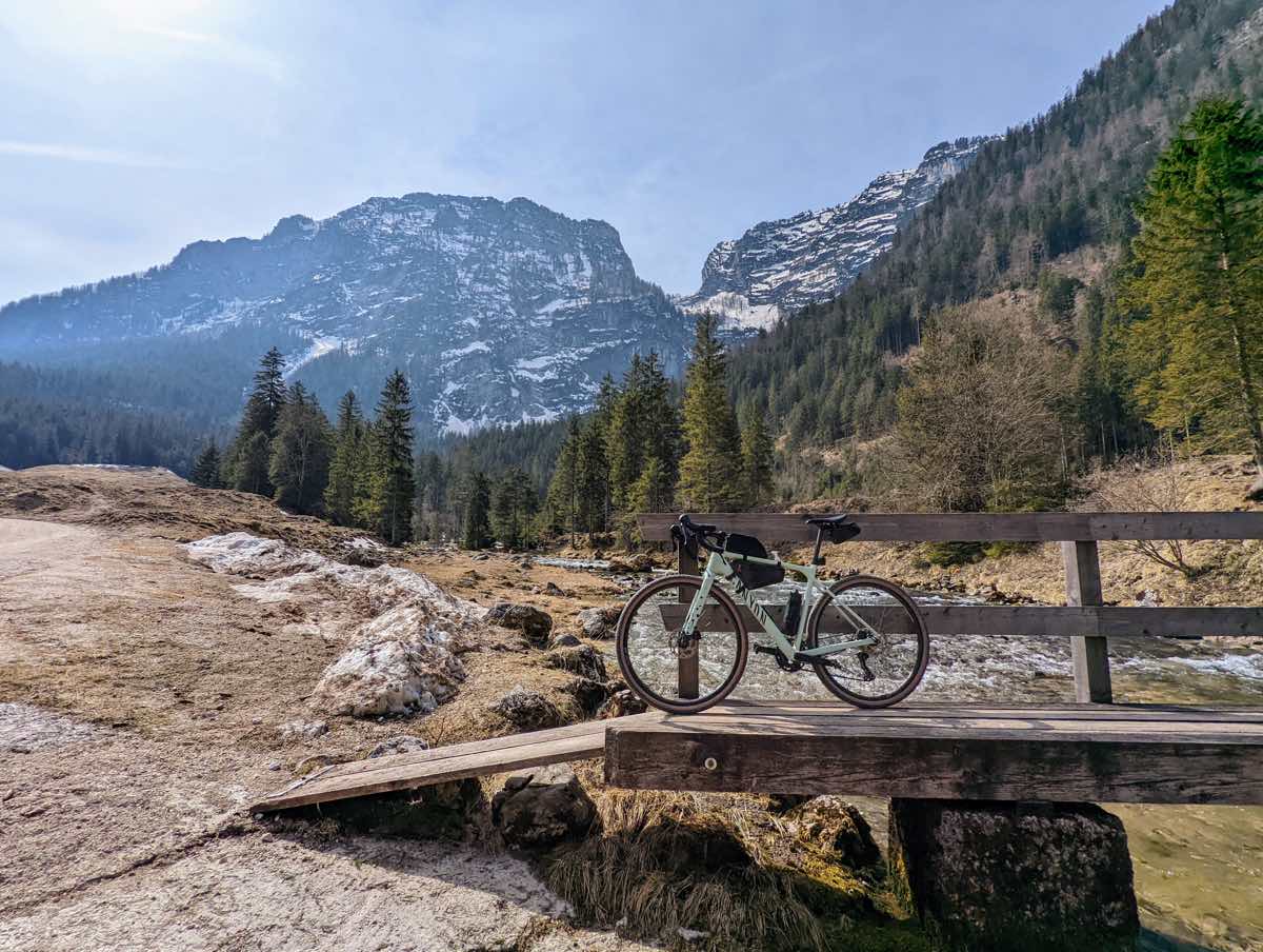 bikerumor pic of the day a bicycle leans against a wood bridge railing, the bridge is spanning a frothy stream that is at the base of a large mountain covered in pine, in the distance is a large rocky mountain covered in patchy snow, the sun is bright.