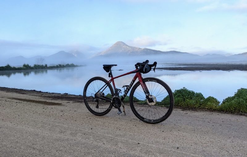 bikerumor pic of the day a bicycle is on a gravel road with a lake beyond it and a small mountain range in the distance surrounded by fog, the sky is clear and bright.