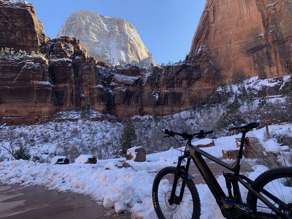 bikerumor pic of the day a emtb is in the foreground of the picture on the side of a road with snow on the ground, the bike is in the bottom of a canyon and looking up are tall rocky cliffs, there is a large light colored cliff in the distance being lit by the sun.