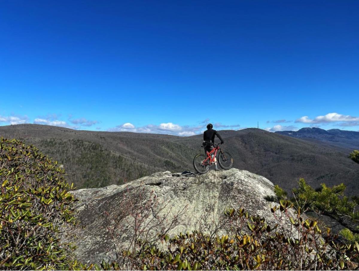bikerumor pic of the day a cyclist is on the top of a high outcropping near the top of a mountain overlooking the surrounding mountains the sky is clear and bright blue