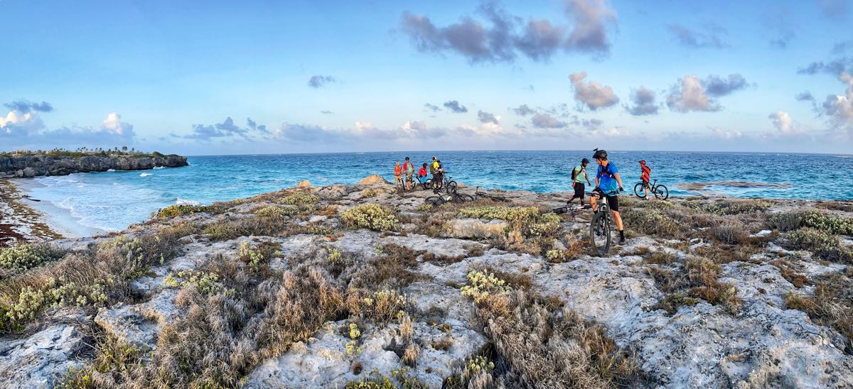 bikerumor pic of the day a group of cyclists explore the rocky point near a beach, the water is clear and there are fluffy puffs of clouds in the sky that mimic the look of the sandy/rocky beach.