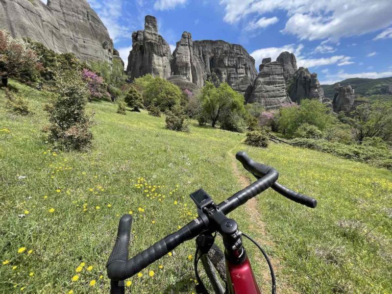 bikerumor pic of the day Meteora, Greece, a photo of a green grassy field leading up to large rocky outcropping front the cockpit of a gravel bicycle.
