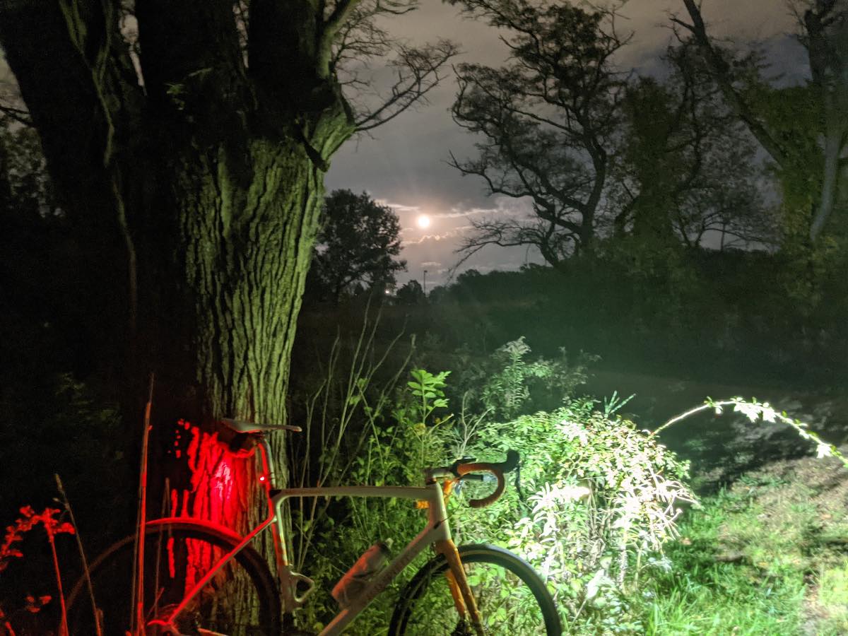 bikerumor pic of the day a bicycle leans agains a large tree trunk at night, it has a front and rear light that are shining on the tree and some bushes, it is night and the moon is shining brightly in the clouds in the distance, the trees and clouds create and eerie photo.