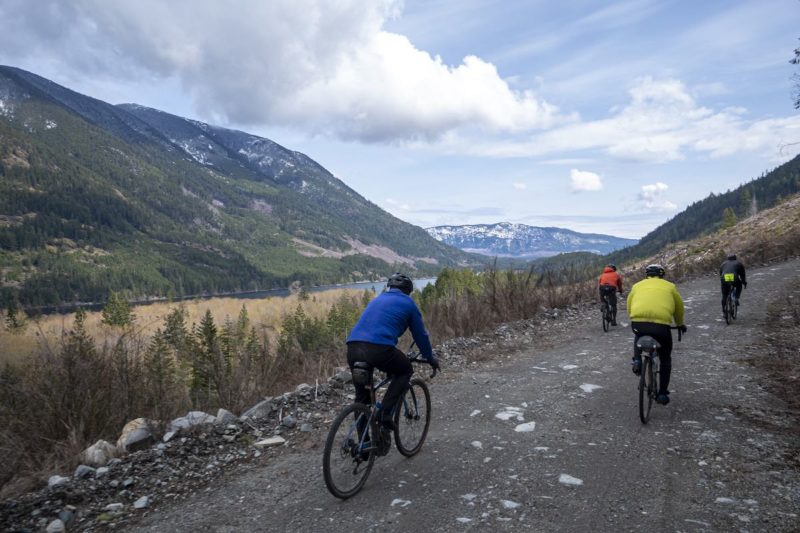 bikerumor pic of the day a group of cyclists riding on a gravel road on the side of a mountain covered in pine trees with a lake down below.