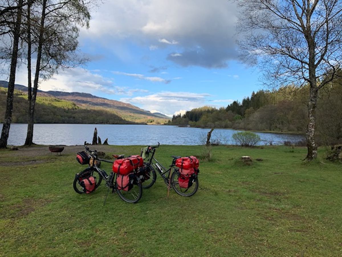 bikerumor pic of the day two touring bicycles loaded with packs are in the grass before a lake in Scotland, the trees surrounding are bare and the sky is spotted with clouds and peeks of blue sky in the distance.