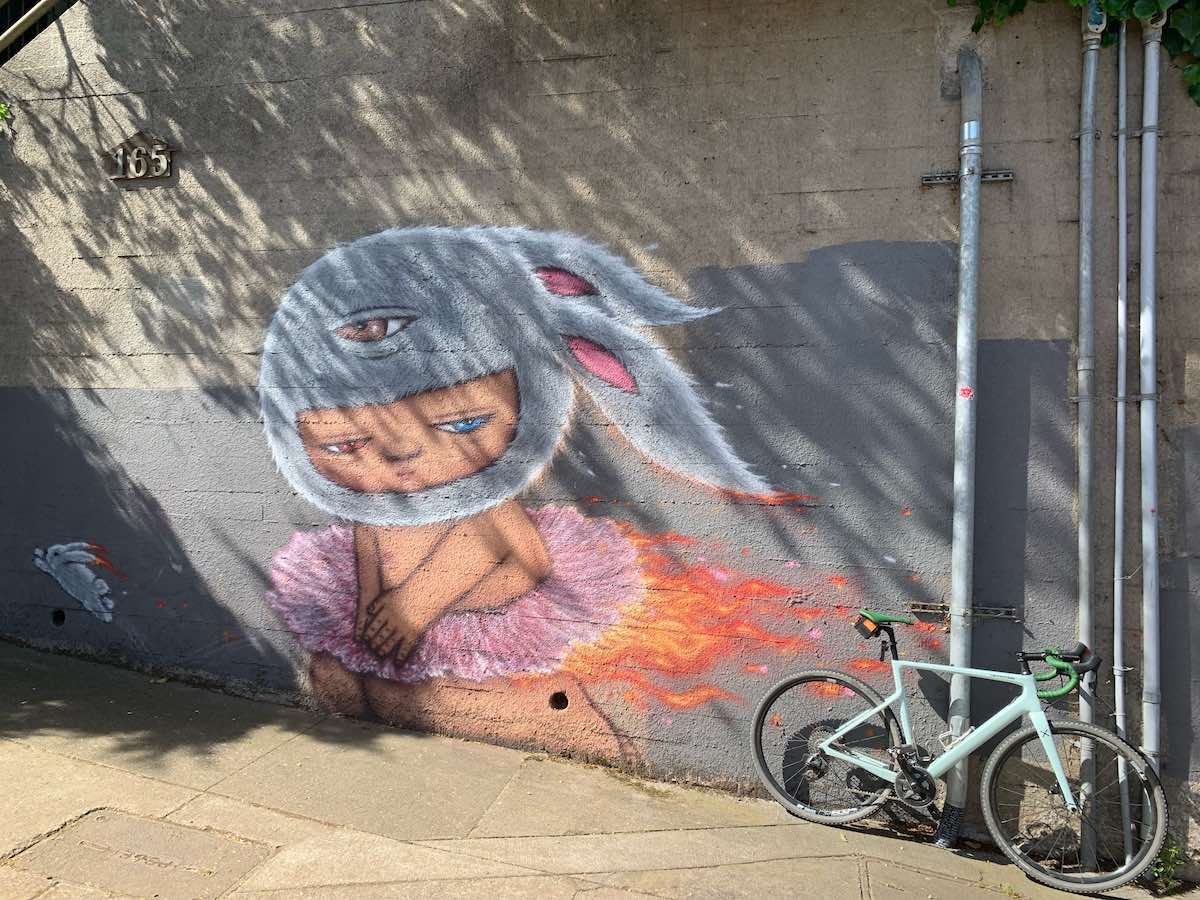 Bikerumor Pic Of The Day, a bicycle is leaning against a wall that has a mural of a child wearing a pink tutu and a bunny hat, the tutu is on fire and the bunny hat has one eye on it.