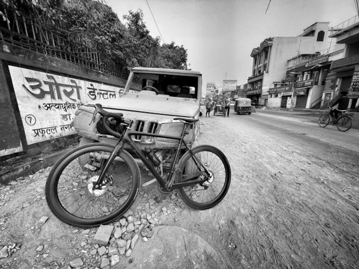 bikerumor pic of the day a gravel bike is leaning against a jeep like car parked next to a concrete wall with indian calligraphy painted on the side, the street is gravel and dirt with chunks of brick near the side, the photo is in black and white.