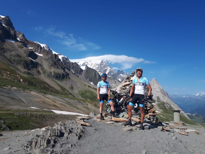 bikerumor pic of the day two gravel cyclists at the top of the col de la Seigne in France, the mountains surrounding are rocky and snow capped and the sun is high and bright.