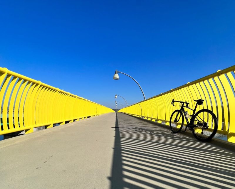 bikerumor pic of the day a bicycle leans against a yellow metal railing along a bridge, the sun is at just the right angle where the shadow of the railing hits the exact center of the bridge, the sky is clear and bright blue.