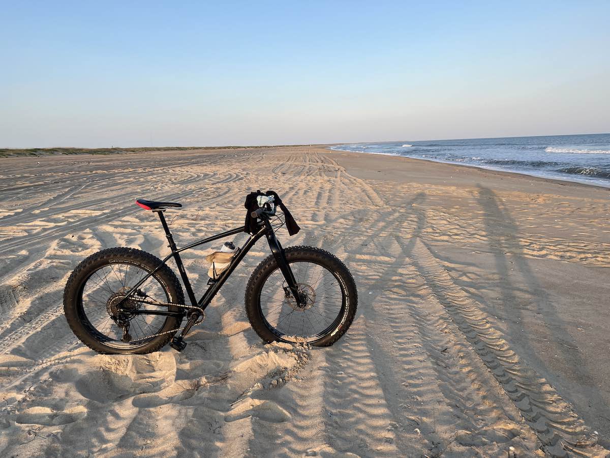bikerumor pic of the day a bicycle with fat tires is poised on a sandy flat beach, the sun is low and setting behind the photographer, the horizon is hazy and pinkish.