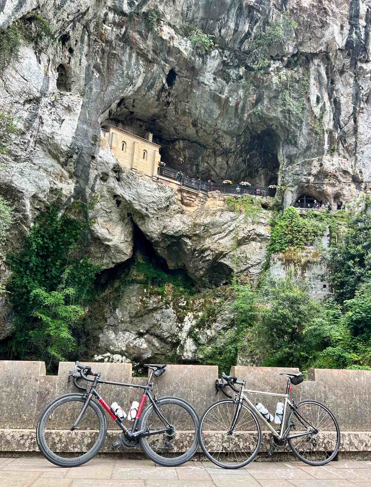 bikerumor pic of the day two bicycles lean against a concrete wall below a cliff with what appears to be a building inside the mouth of a cave.